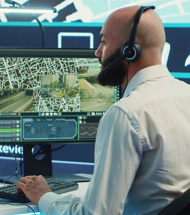 Un hombre barbudo con auriculares supervisa los mapas de la ciudad y las imágenes de las cámaras de seguridad en la pantalla de un ordenador en una sala de control.