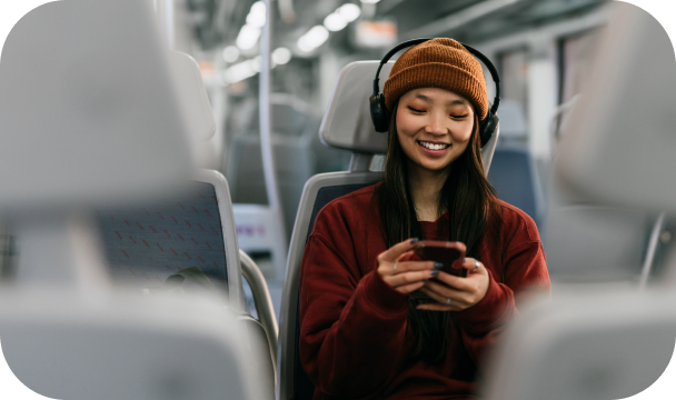 En ung kvinne sitter på et tog og smiler. Hun har på hodetelefoner og holder mobiltelefonen i begge hender, noe som kan tyde på at hun jobber på vei hjem.