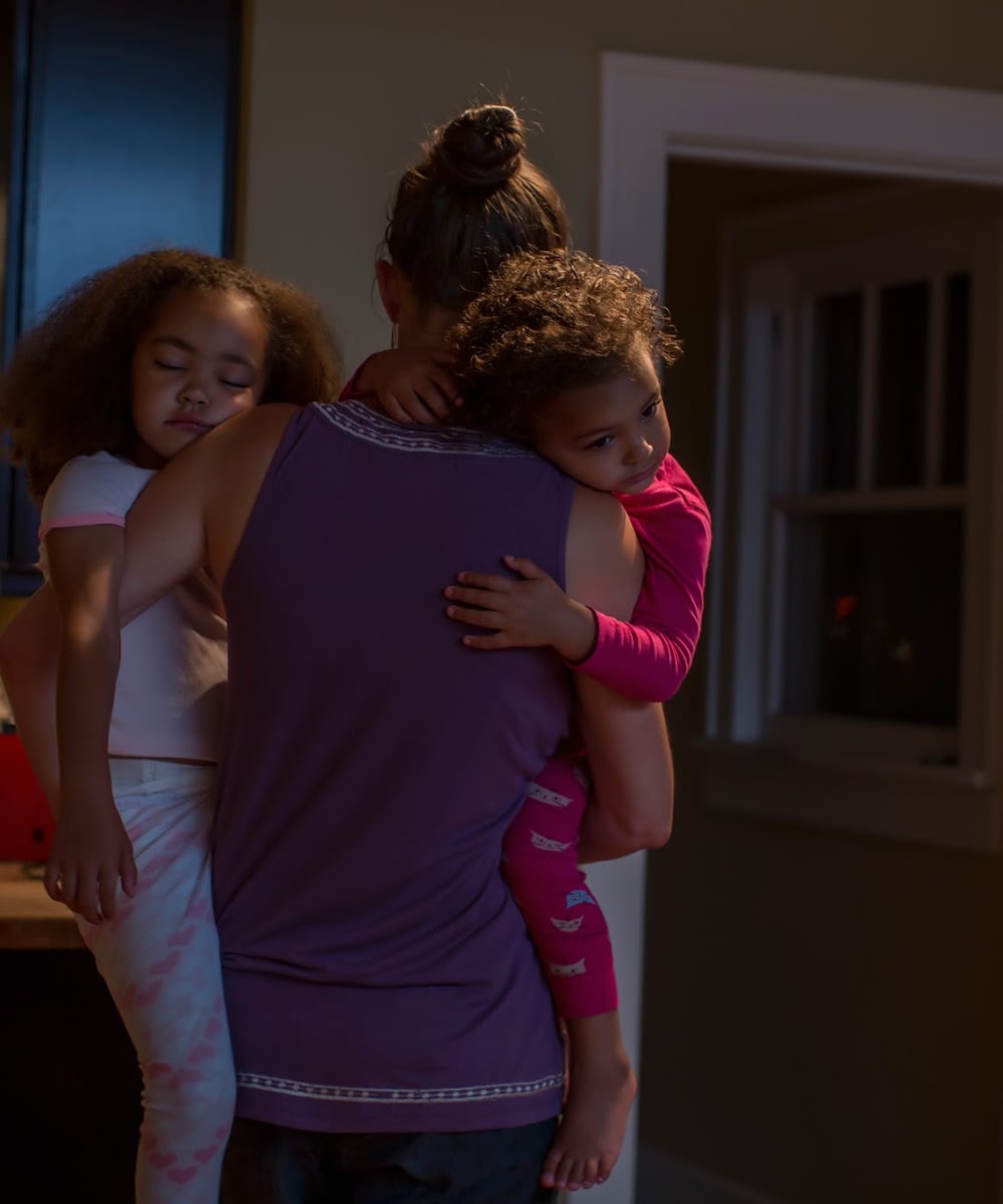 an image of a woman carrying in her arms her two daughters falling asleep to their bedrooms